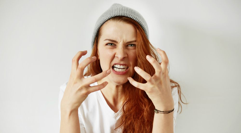 A woman wearing a gray beanie is making an aggressive gesture with her hands and baring her teeth in a snarling expression, indicating strong emotions like anger or frustration. The background is plain and light, focusing attention on her expressive face.