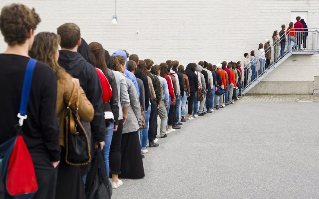 A long line of people is seen queuing up against a blank wall, extending back towards a staircase where more individuals are gathered. The diversity in clothing suggests it could be a public or social event. Everyone is facing in one direction, indicative of waiting for entry or service.