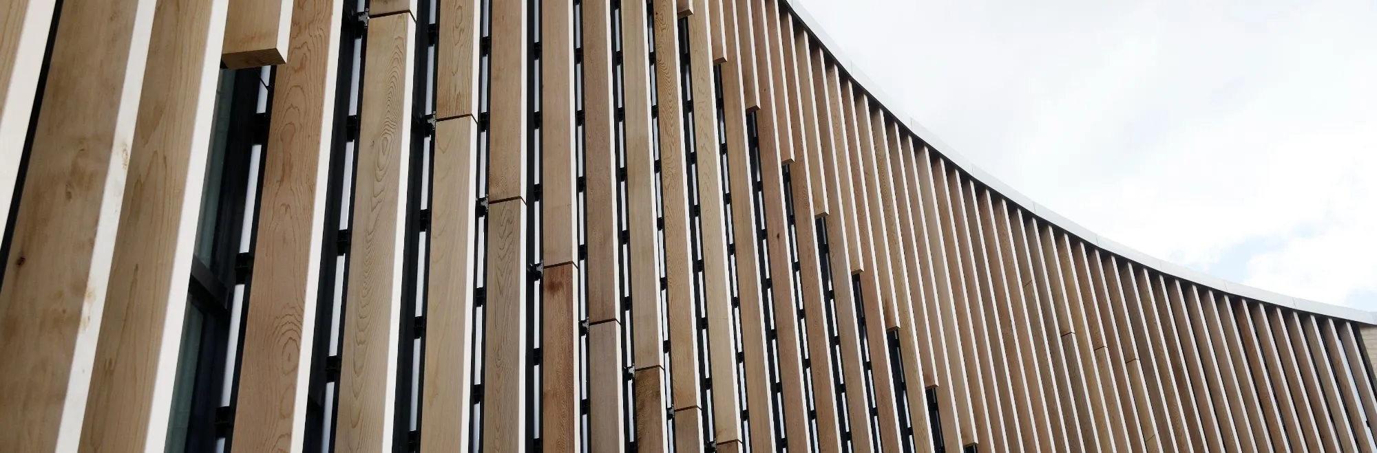 Moderne Fassade mit vertikal angeordneten Holzelementen und Metallstreben vor wolkigem Himmel.