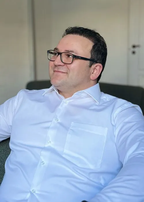 Mann mit schwarzer Brille und weißem Hemd sitzt entspannt und schaut lächelnd zur Seite.