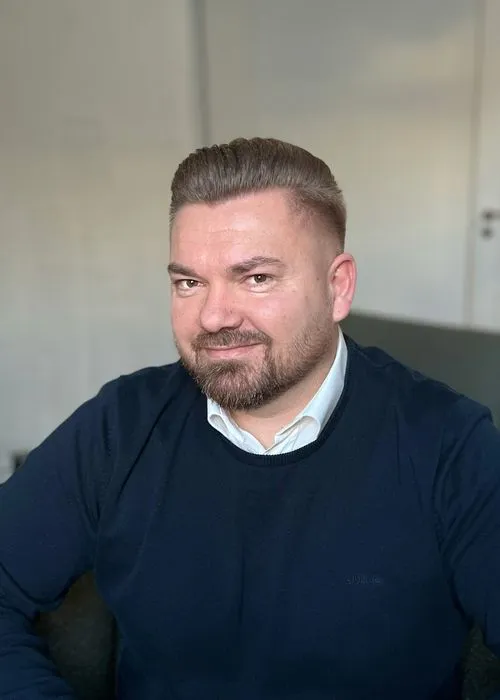 Porträt eines Mannes mit kurzem braunem Haar und Bart, der einen weißen Kragen und einen dunkelblauen Pullover trägt.
