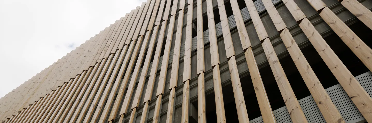 Blick nach oben auf eine Gebäudefassade mit vertikal angeordneten hellen Holzlatten vor einer Betonwand.