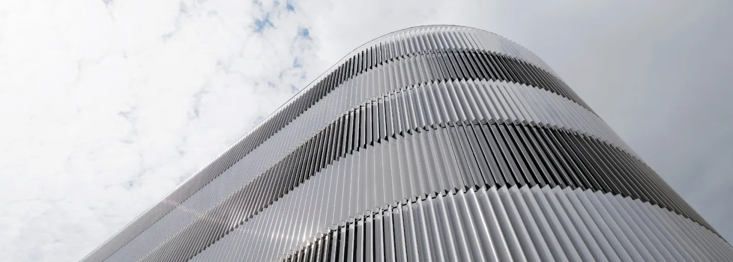 Blick nach oben auf ein modernes, geschwungenes Hochhaus mit vertikalen Lamellen und bewölktem Himmel im Hintergrund.