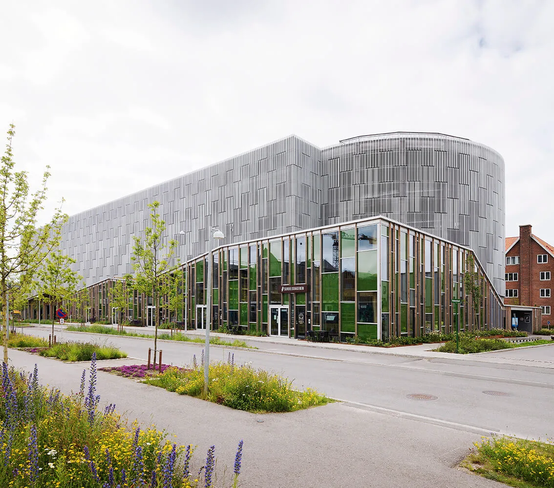 Modernes Bürogebäude mit großen Glasfenstern, grünen Pflanzen und einer Fassade aus vertikalen Metallstreifen.