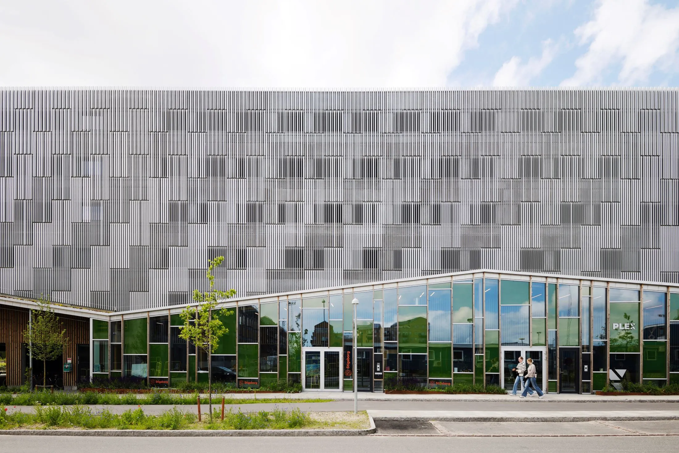 Modernes Gebäude mit einer Fassade aus vertikalen weißen Lamellen und großen Glasfenstern im unteren Bereich, davor zwei Menschen, die auf dem Bürgersteig gehen.