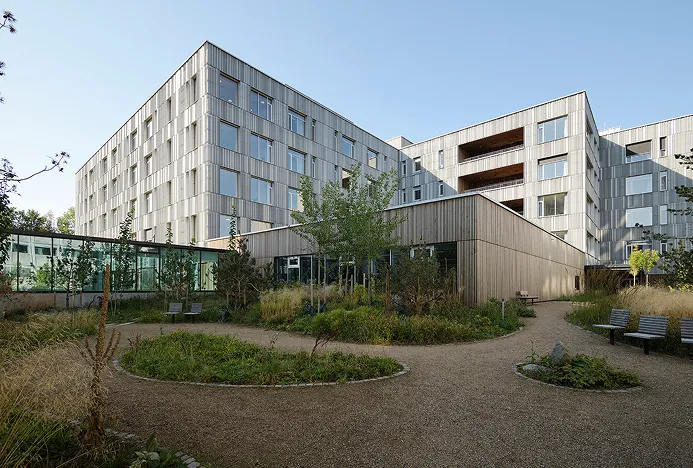 Modernes Krankenhausgebäude mit mehreren Etagen umgeben von einem Garten mit Bänken und Kieswegen.