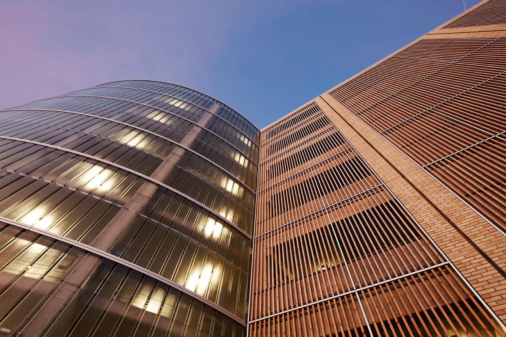 Blick nach oben auf zwei moderne Gebäude mit Glas- und Holzlamellenfassaden unter blauem Himmel.