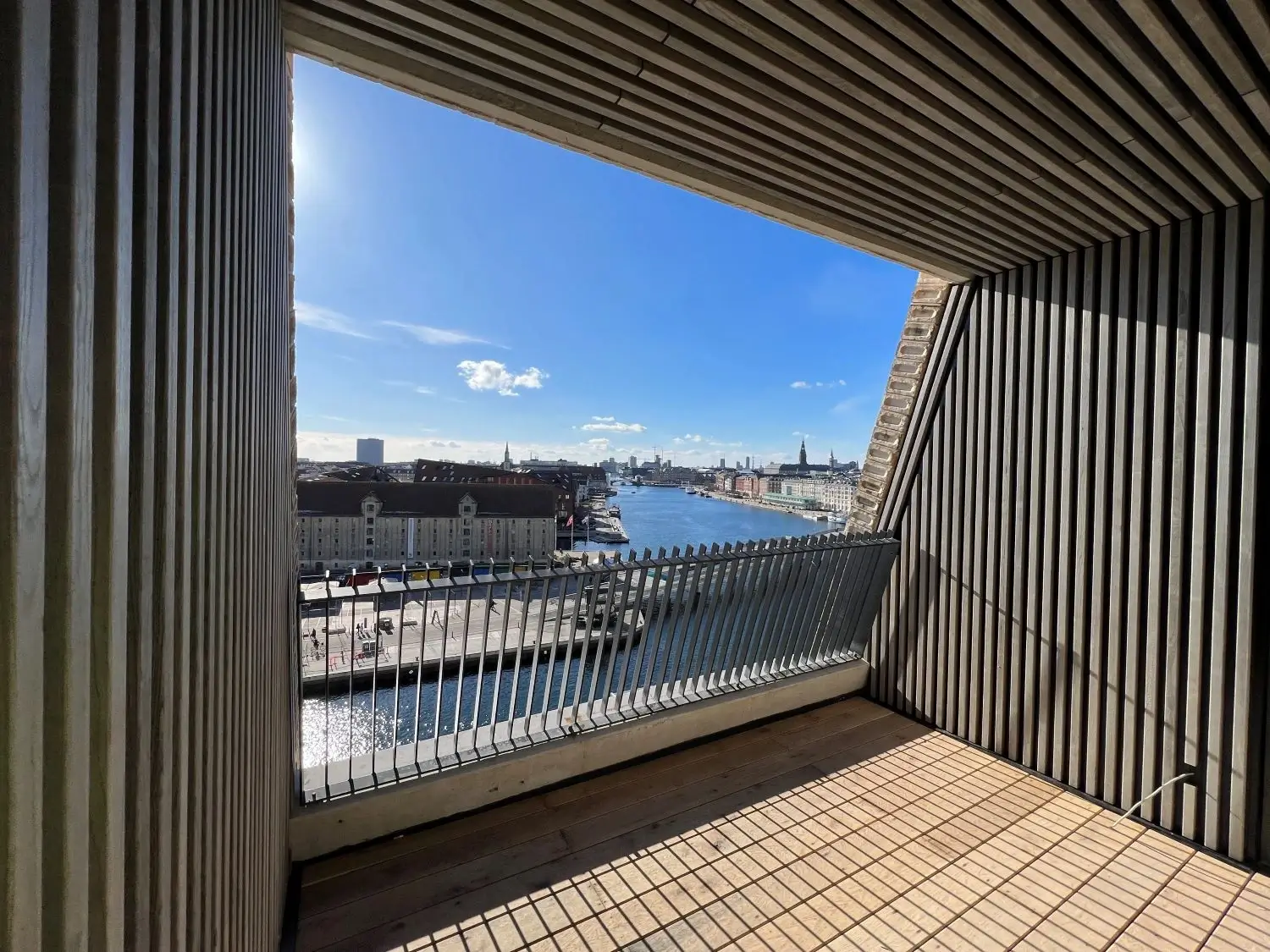 Blick auf einen Kanal und Gebäude in einer Stadt von einem Balkon mit Holzverkleidung und Metallgeländer an einem sonnigen Tag.