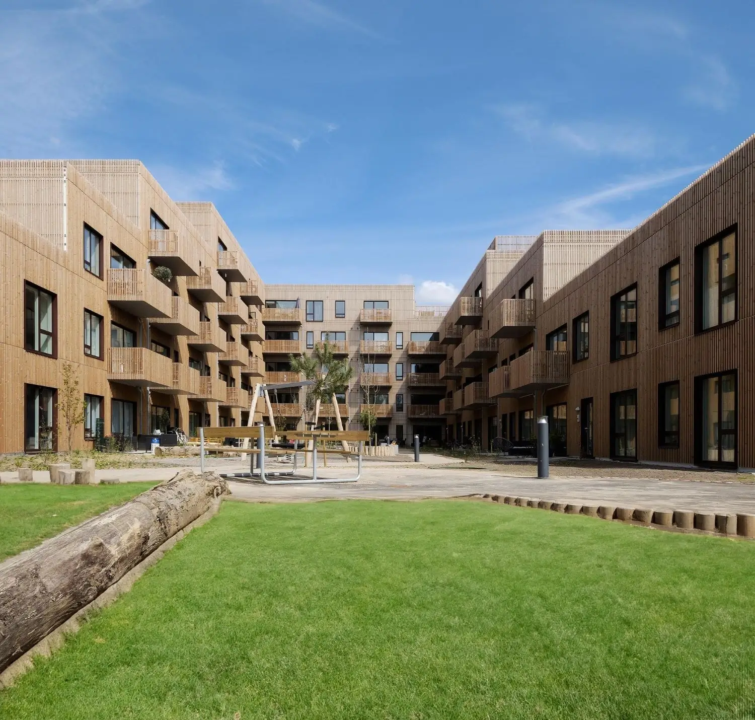 Innenhof eines Wohngebäudes mit Holzfassaden, kleinen Balkonen, grüner Rasenfläche und Spielplatz unter blauem Himmel.