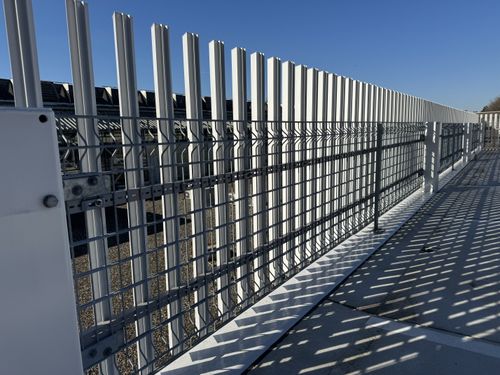 Metalldrahtzaun und vertikale weiße Stahlträger mit Schatten auf Betonboden unter klarem blauem Himmel.