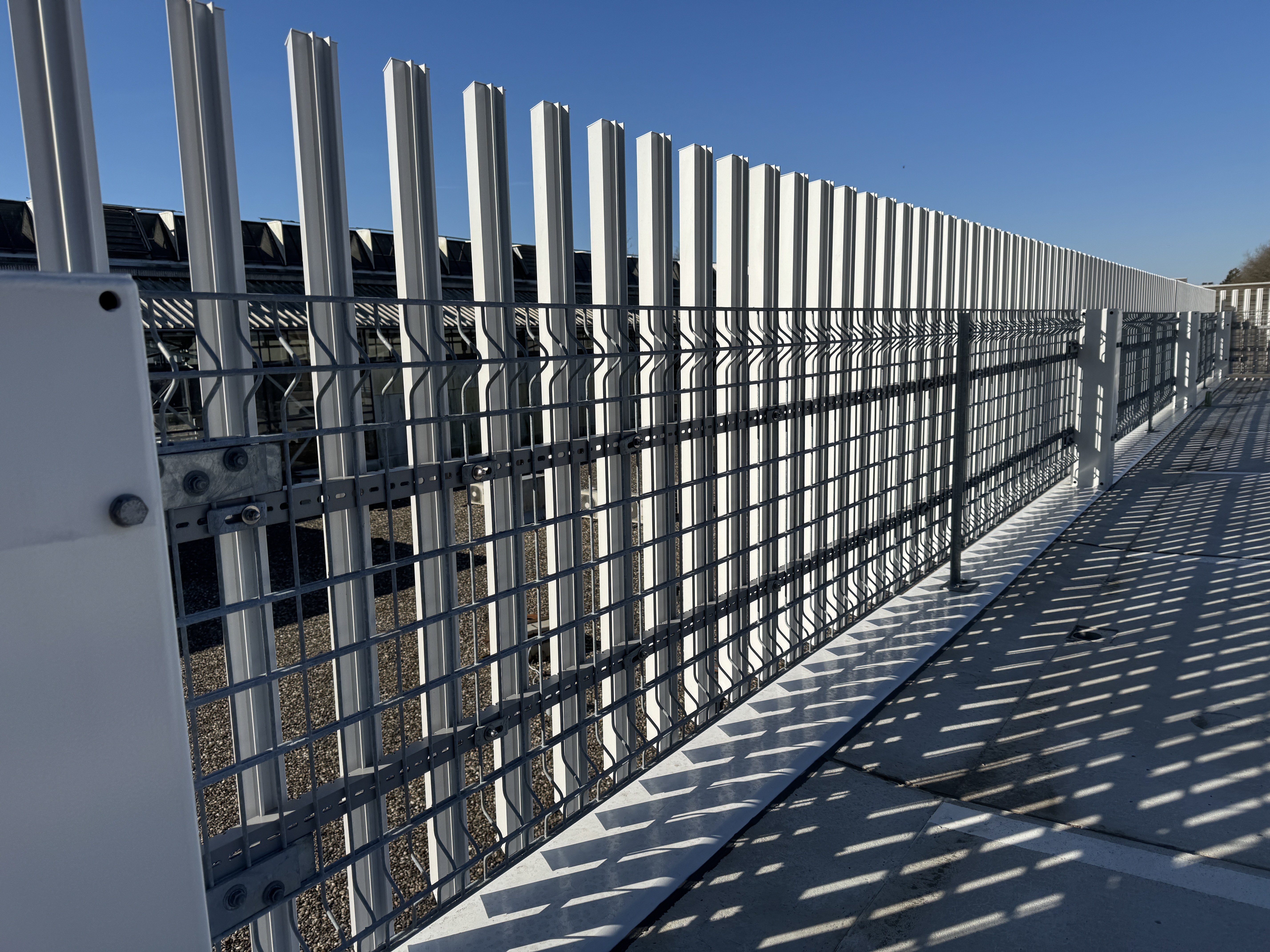 Metalldrahtzaun und vertikale weiße Stahlträger mit Schatten auf Betonboden unter klarem blauem Himmel.