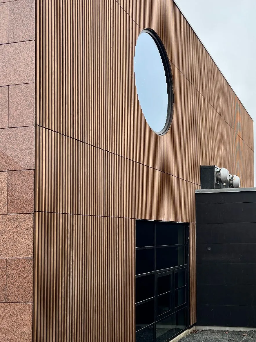 Fassade aus vertikalen Holzlamellen mit rundem Fenster neben dunklem Gebäudeteil