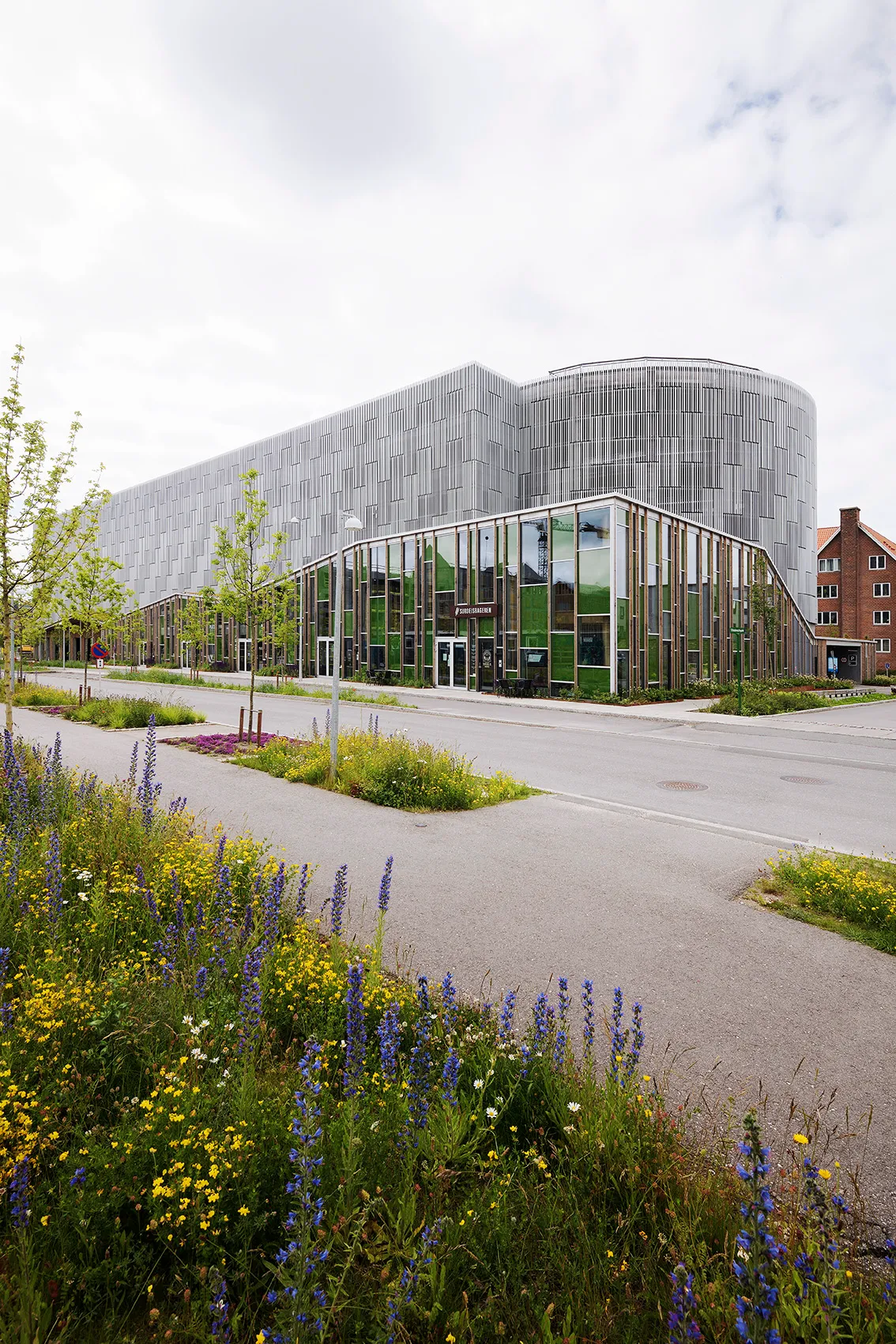 Stigsborg mit gerundetem Parkhaus und Lamellenfassade hinter Wildblumenwiese, Woodfac Click und Napsu