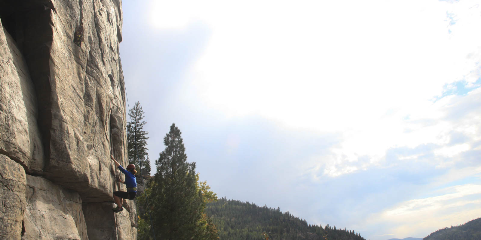 A breathtaking view of a rock climber mid-route, hanging above a ledge, with a mountain and forest background.