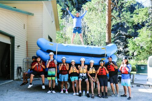 A raft guide standing on top of a raft pile while a group of boys stand beneath, posing for a photo.