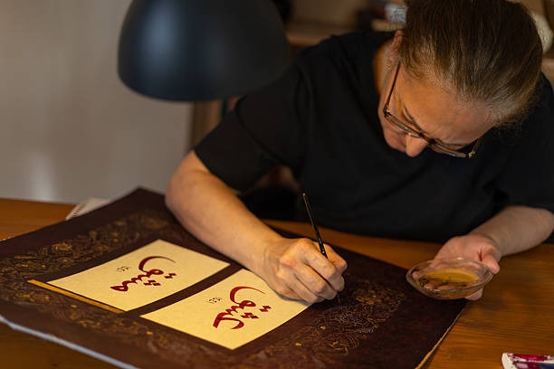 A woman practicing Arabic calligraphy art at a wooden desk, using traditional ink and brush techniques. The artwork features intricate script on parchment paper with decorative borders. This image highlights Emirati cultural workshops, perfect for UAE National Day office activities, team-building experiences, or events focused on Arabic heritage and traditional arts in the UAE.