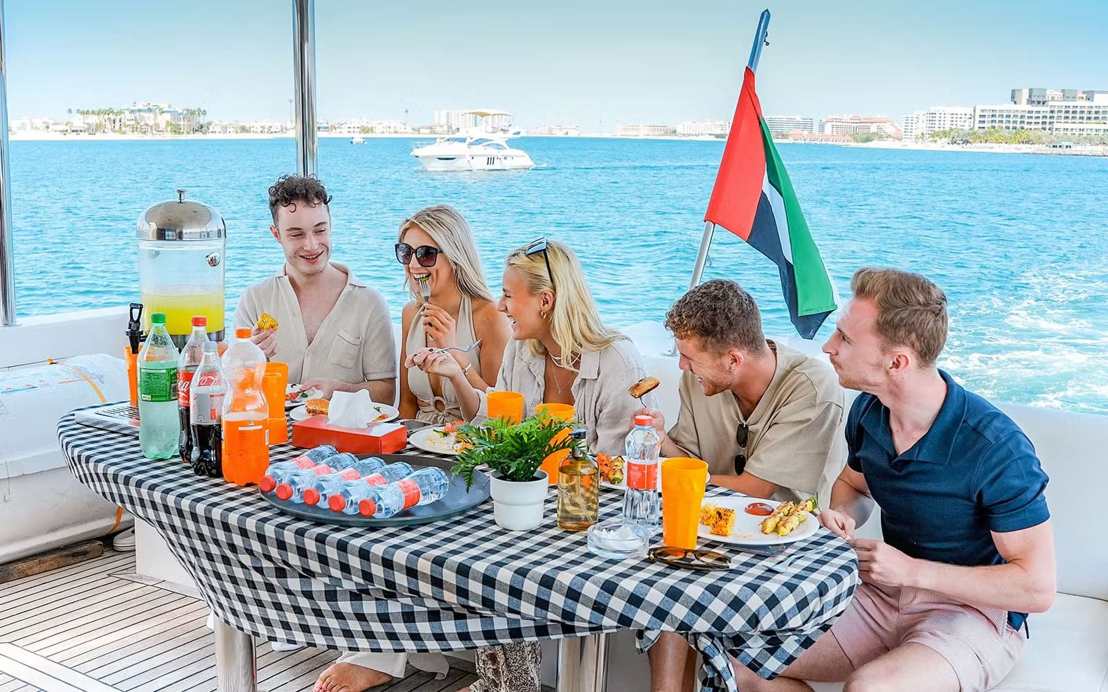 A group of young professionals enjoying a UAE National Day yacht party in Dubai. The group is gathered around a checkered table filled with food, soft drinks, and decorations, with the UAE flag waving in the background. Set against the clear blue waters and city skyline, this team celebration on a private yacht showcases a premium and fun way to celebrate National Day in Dubai with colleagues or friends.