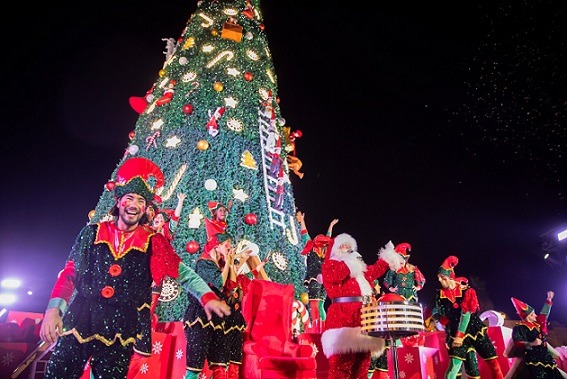 A vibrant Christmas celebration featuring Santa Claus and cheerful elves performing in front of a giant, beautifully decorated Christmas tree at a public event. The scene is lit with festive lights and surrounded by gift boxes, capturing the joyful spirit of holiday parades, Christmas tree lightings, or family-friendly Christmas events in Dubai.