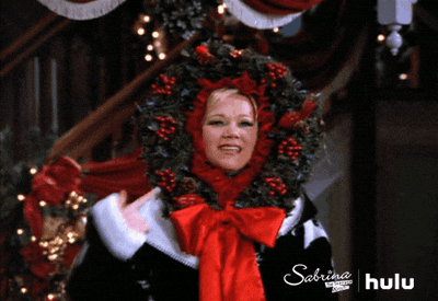 Woman dressed as a Christmas wreath with red bows and holly, smiling joyfully in a festive holiday scene from Sabrina the Teenage Witch on Hulu.