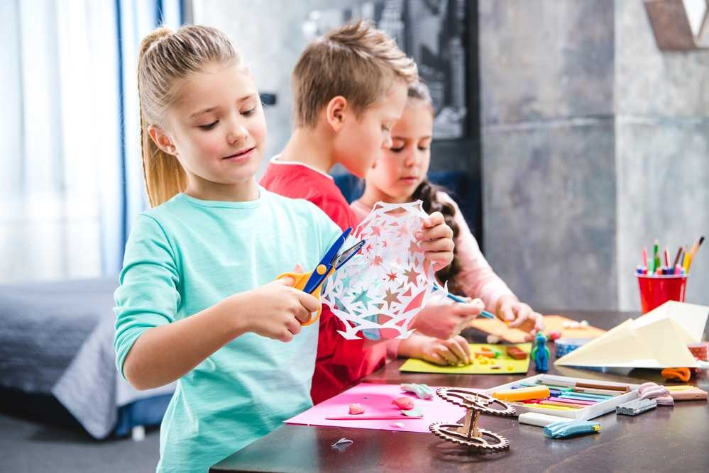 Three children focused on arts and crafts, cutting paper snowflakes and creating colorful projects with markers, clay, and paper at a craft table.