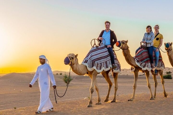 Family riding camels during a desert safari at sunset in Dubai, guided by a local Bedouin in traditional dress.