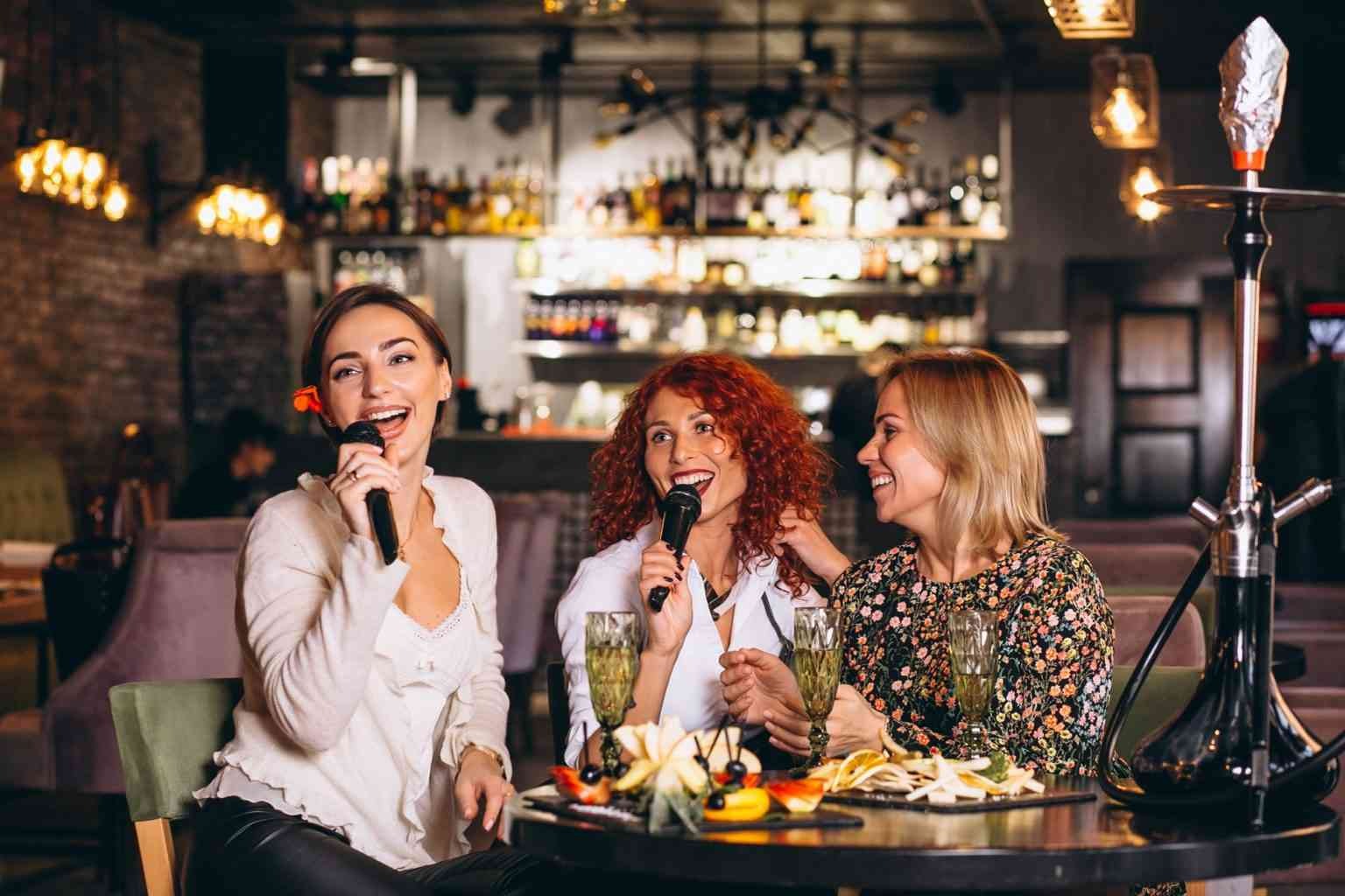 Three women singing enthusiastically at a karaoke night in Dubai, surrounded by cheering friends.