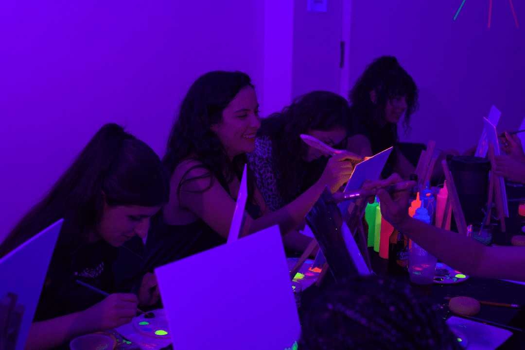 Friends laughing and painting with neon colors under blacklight at a UV art session in Dubai.