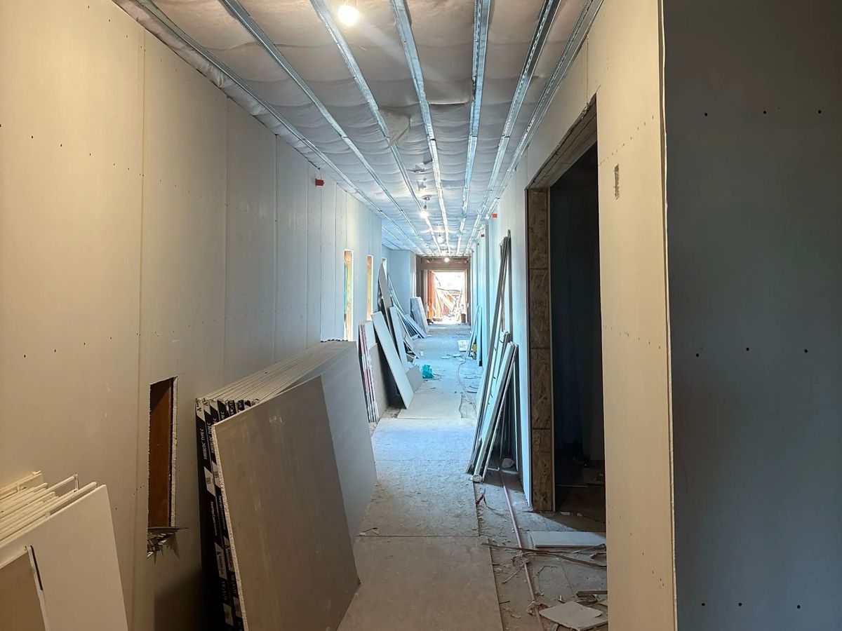 Multifamily corridor during drywall installation, with drywall sheets leaning against hung board walls and metal ceiling framing overhead