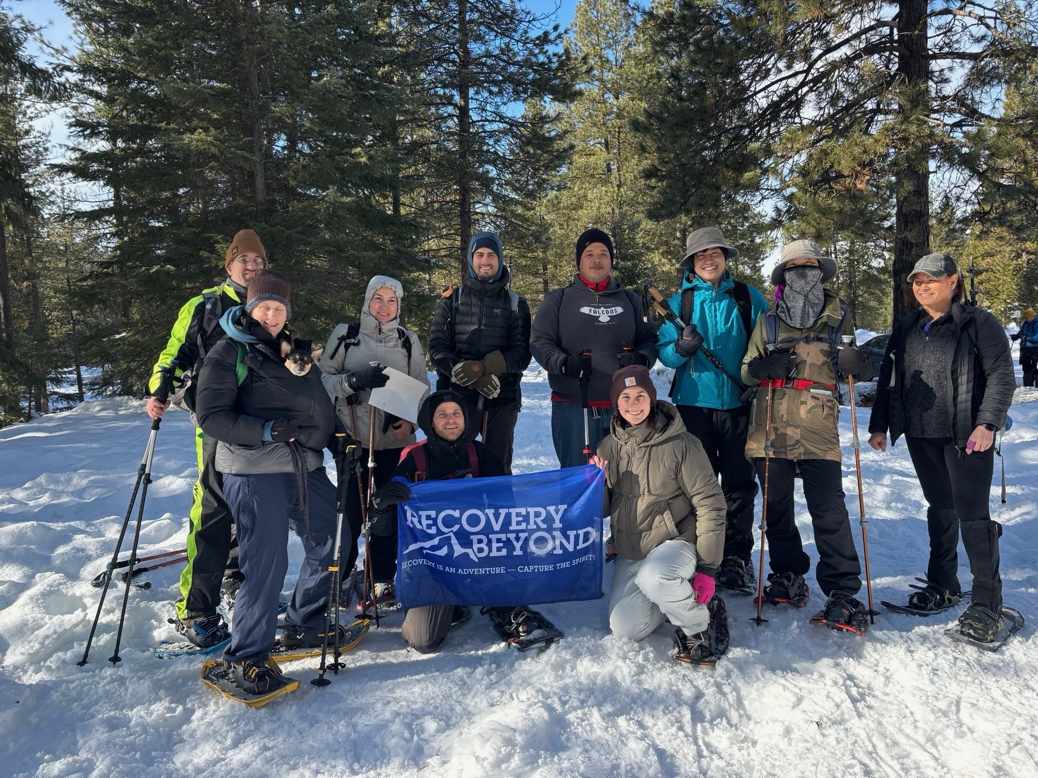 Group of people wearing winter clothing and snowshoes standing and kneeling in snow surrounded by trees, holding a blue banner reading 'Recovery Beyond'.