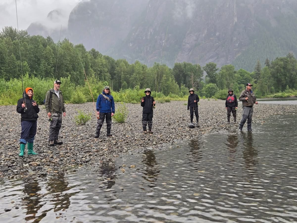 Seven people standing on a rocky riverbank with fishing rods, surrounded by trees and misty mountains.