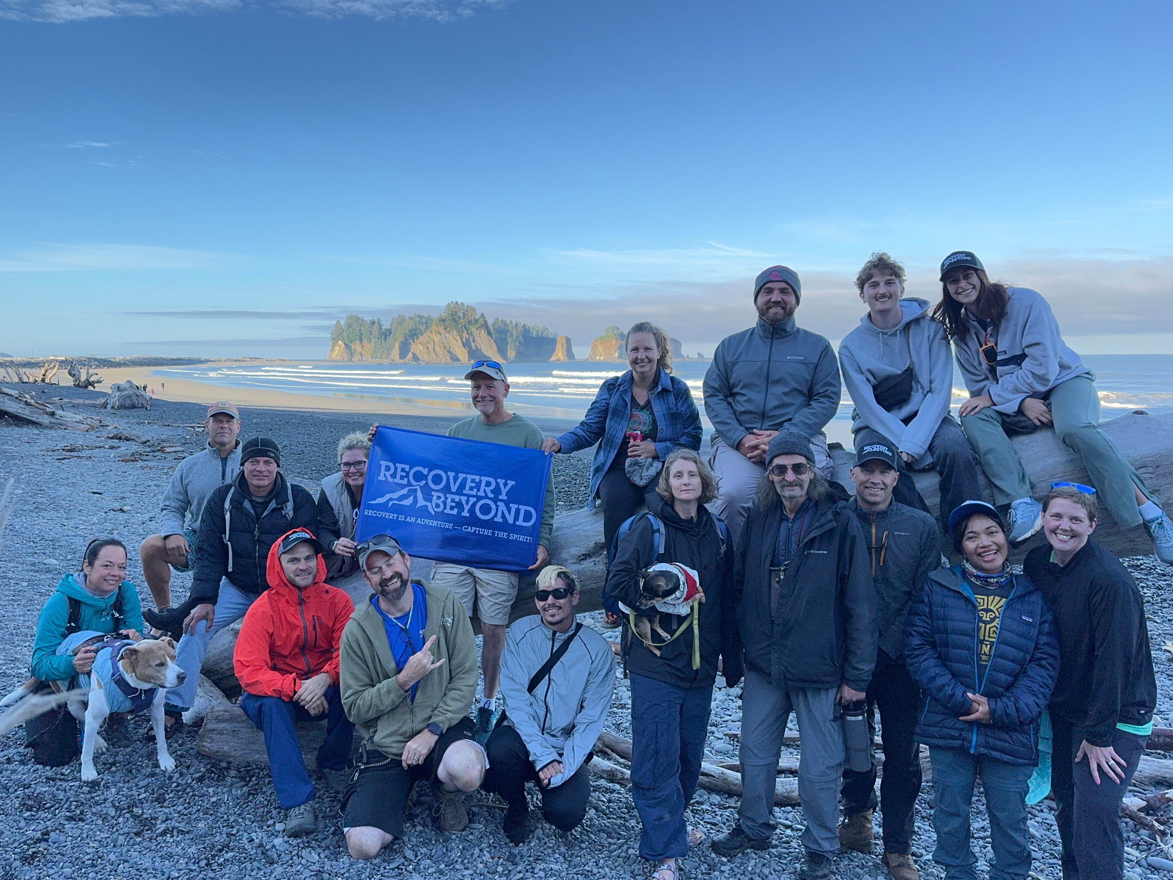 Group of people posing on a rocky beach near ocean with driftwood and island rocks in background, one holding a blue 'Recovery Beyond' banner.
