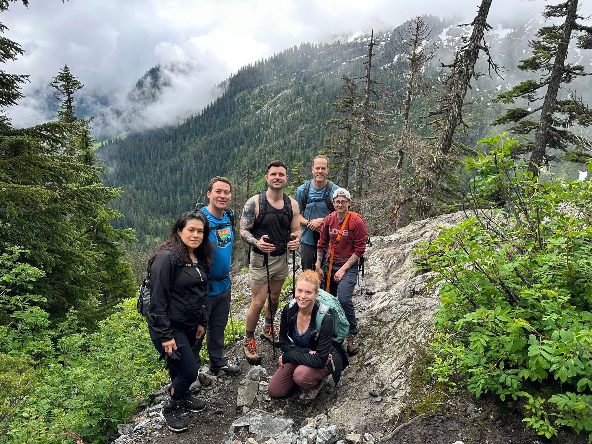 Group of six hikers posing on a rocky mountain trail surrounded by evergreen trees and misty peaks.