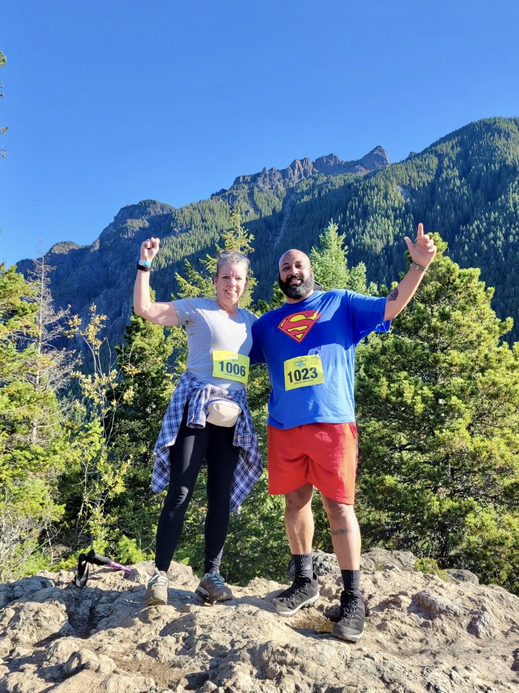 Two hikers with race bibs 1006 and 1023 posing triumphantly on rocky terrain with forested mountains and clear blue sky in the background.