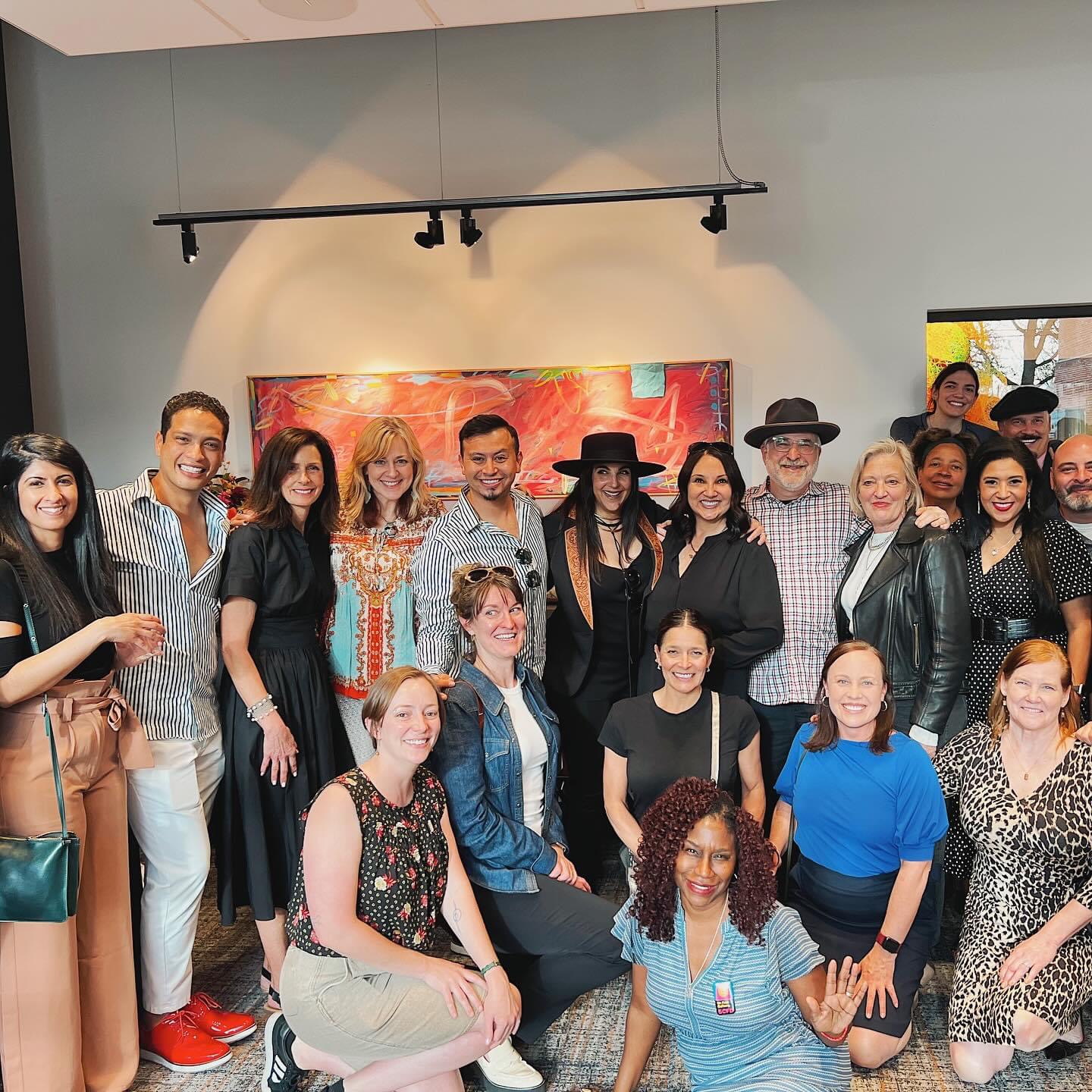 20 people of all ages and descriptions pose for a photo, a painting is in the background