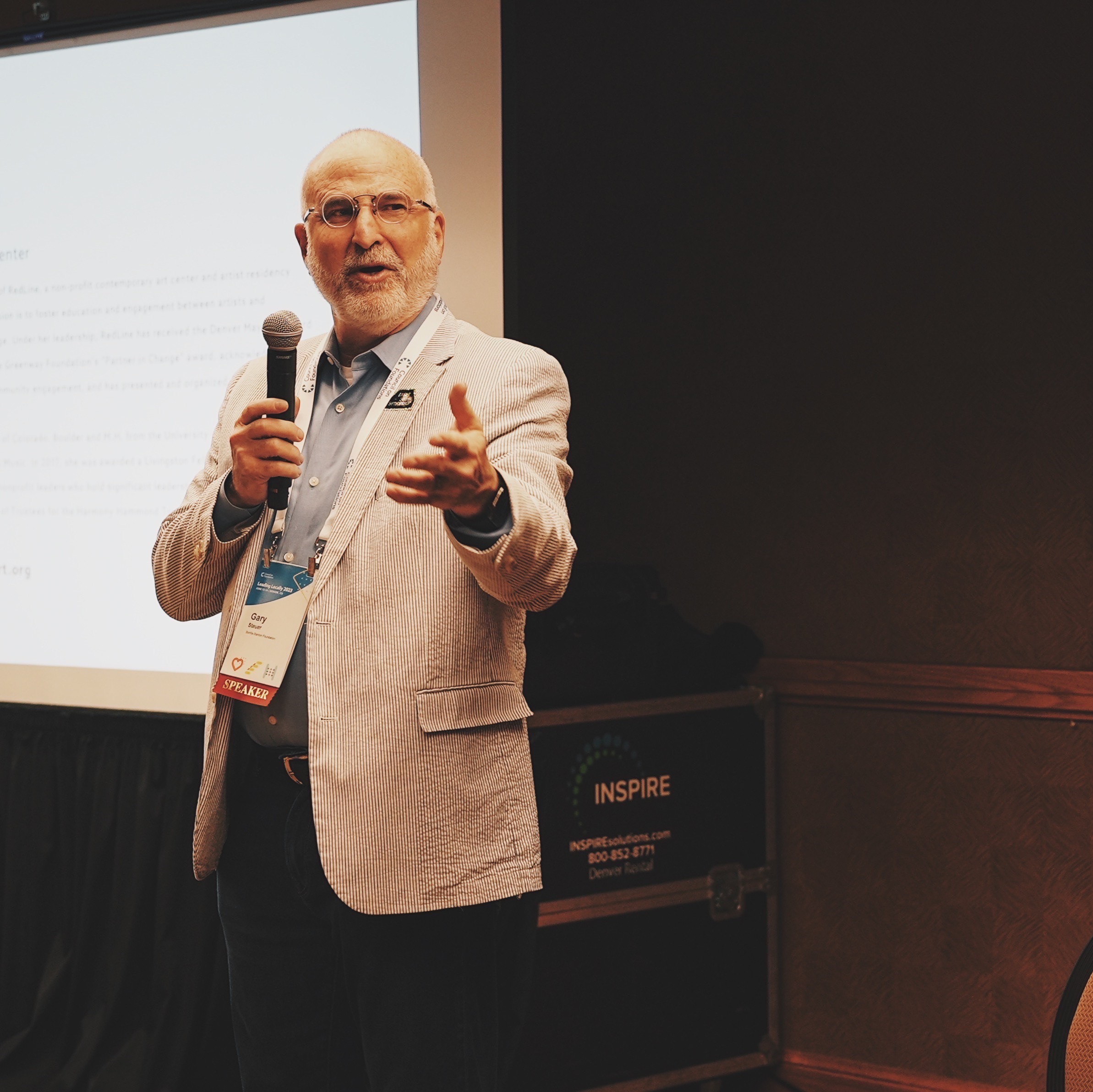 Gary Steuer is a white man with a bald head and white beard, wearing glasses, a seersucker jacket, and speaking into a microphone in front of a projection screen 