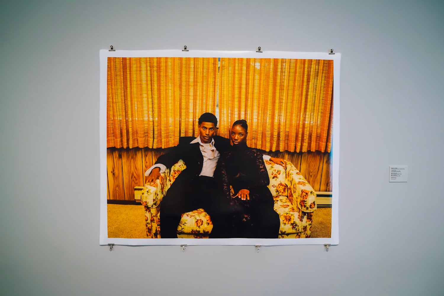 A framed portrait by Marcus Correa showing two figures seated against gold curtains