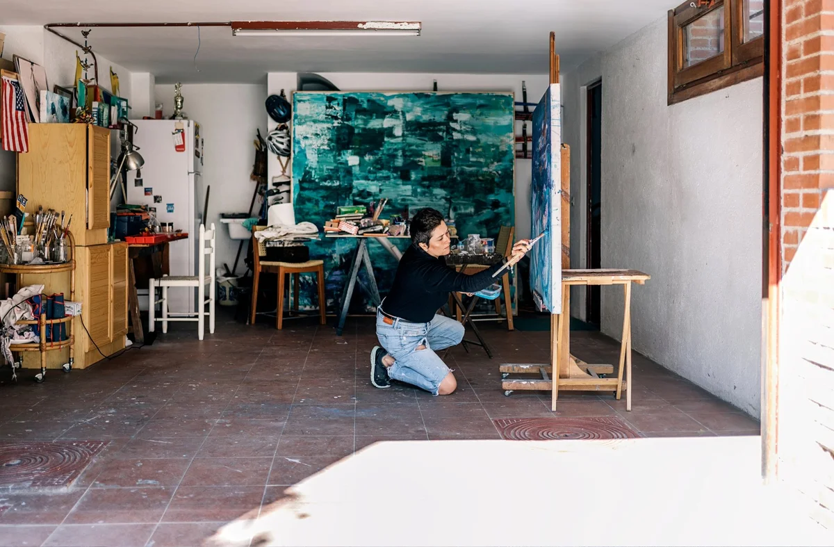 Artist kneeling and painting on a canvas in a spacious, well-lit studio with art supplies.