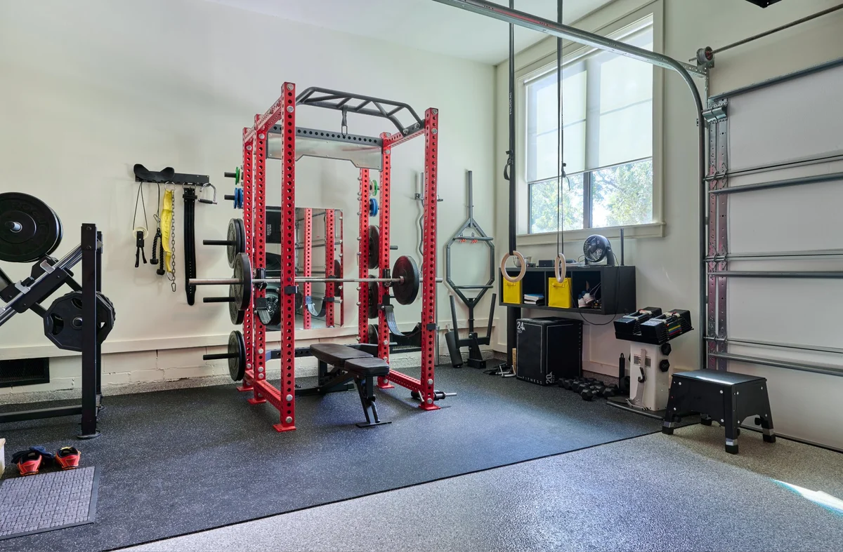 Home gym with red power rack, bench, dumbbells, gymnastic rings, weighted plates, and exercise equipment.