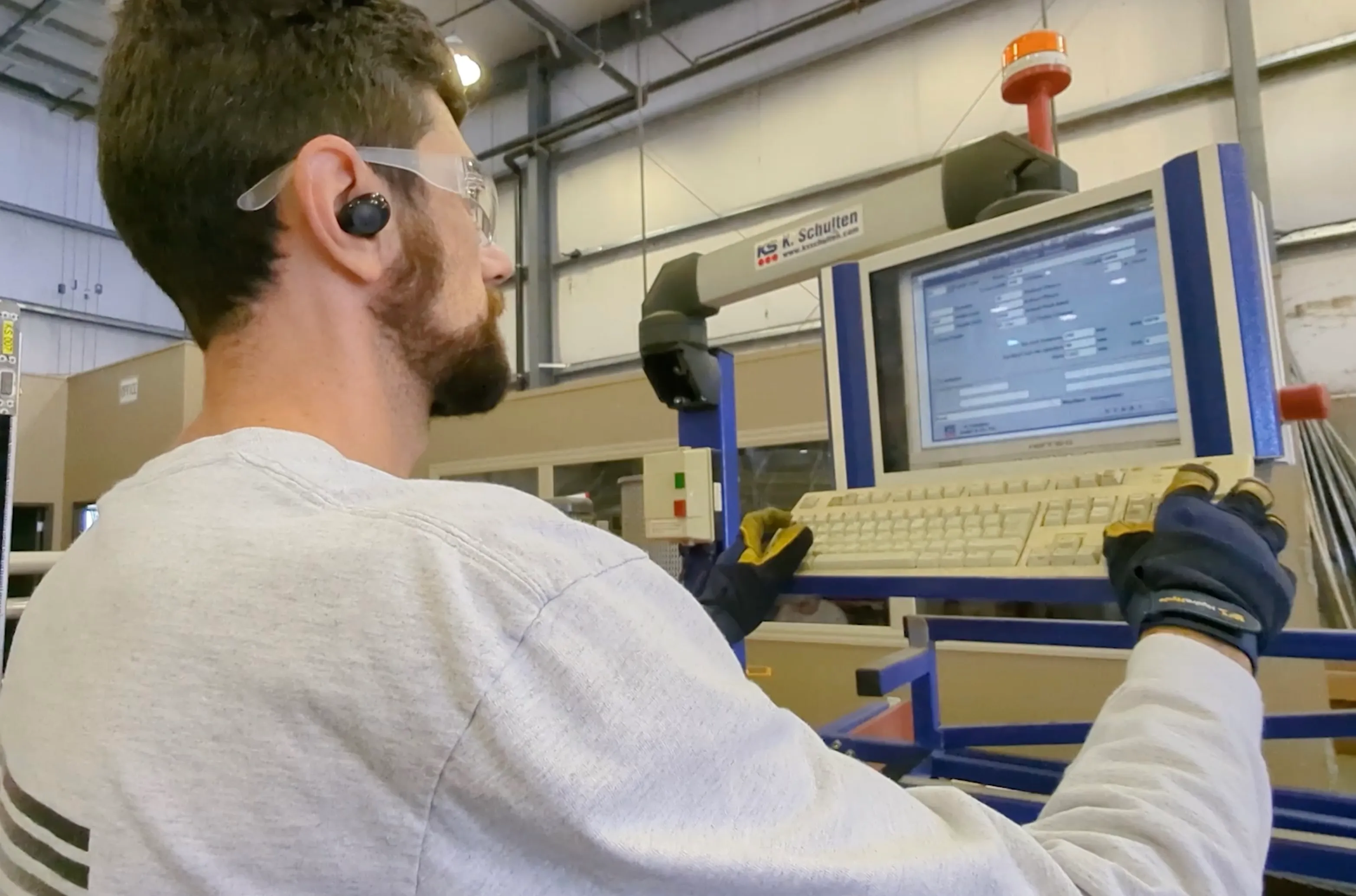 Man wearing safety glasses and gloves operates a machine control panel with keyboard in industrial setting.