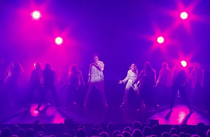 Konsertscen med två artister i vita kostymer under lila scenbelysning