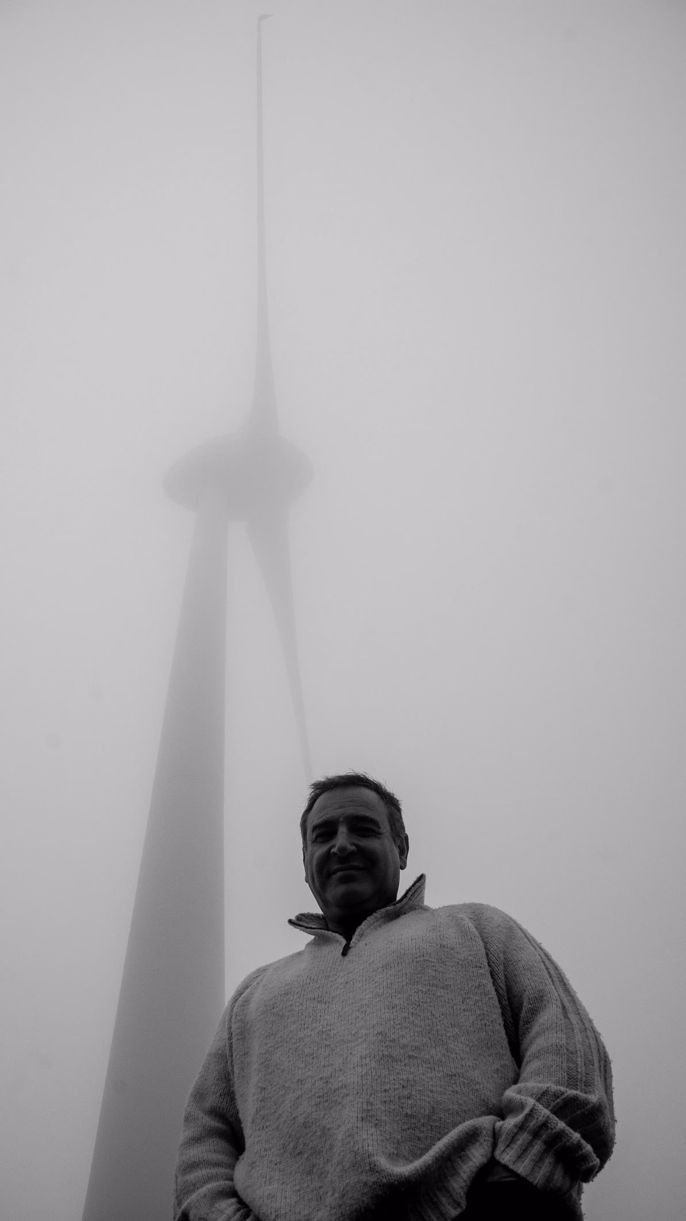 Black and white photo of John Psathas smiling in a knit sweater below a wind turbine disappearing into fog