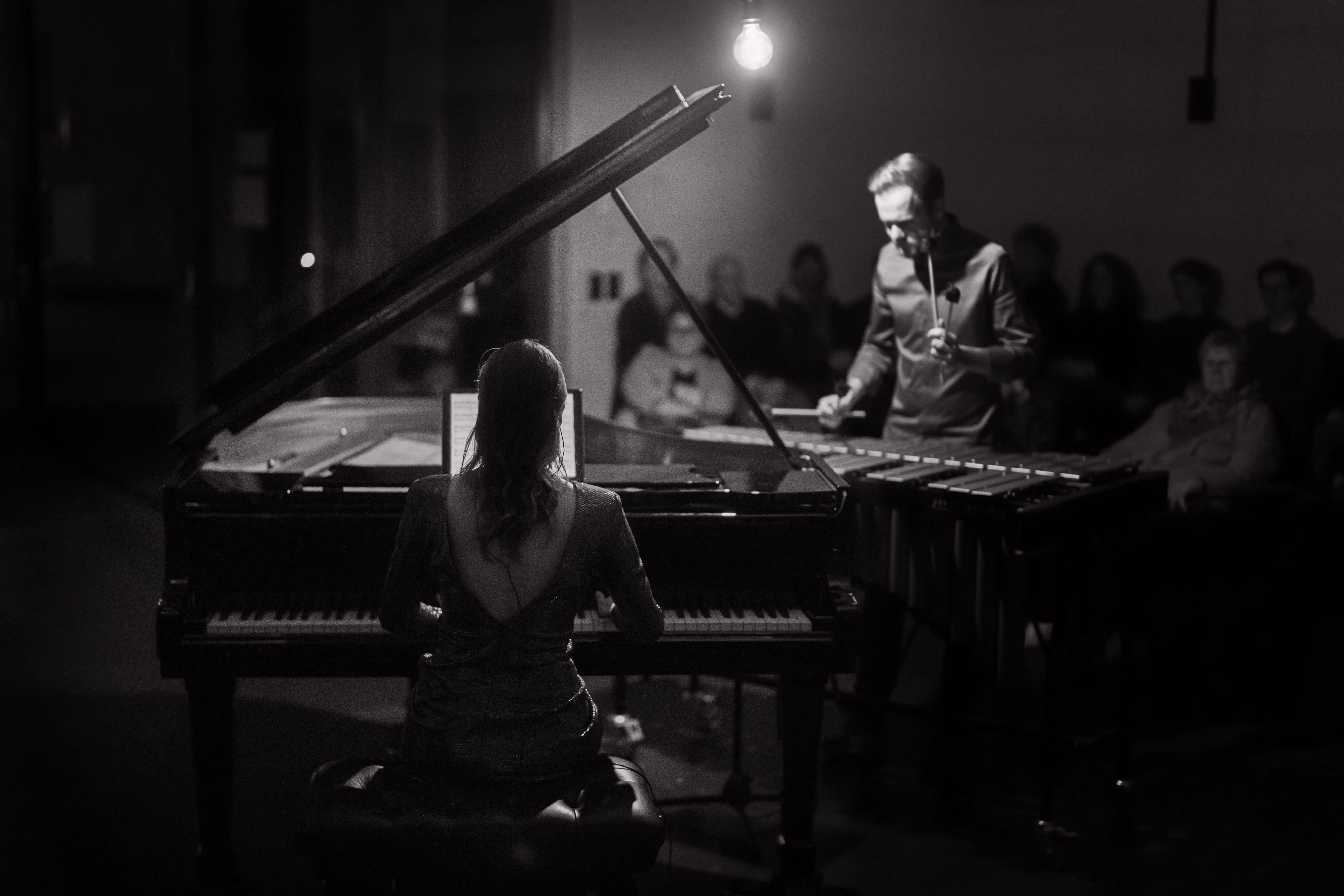 Black and white concert photo of a pianist at a grand piano and a percussionist on marimba performing John Psathas music for a seated audience