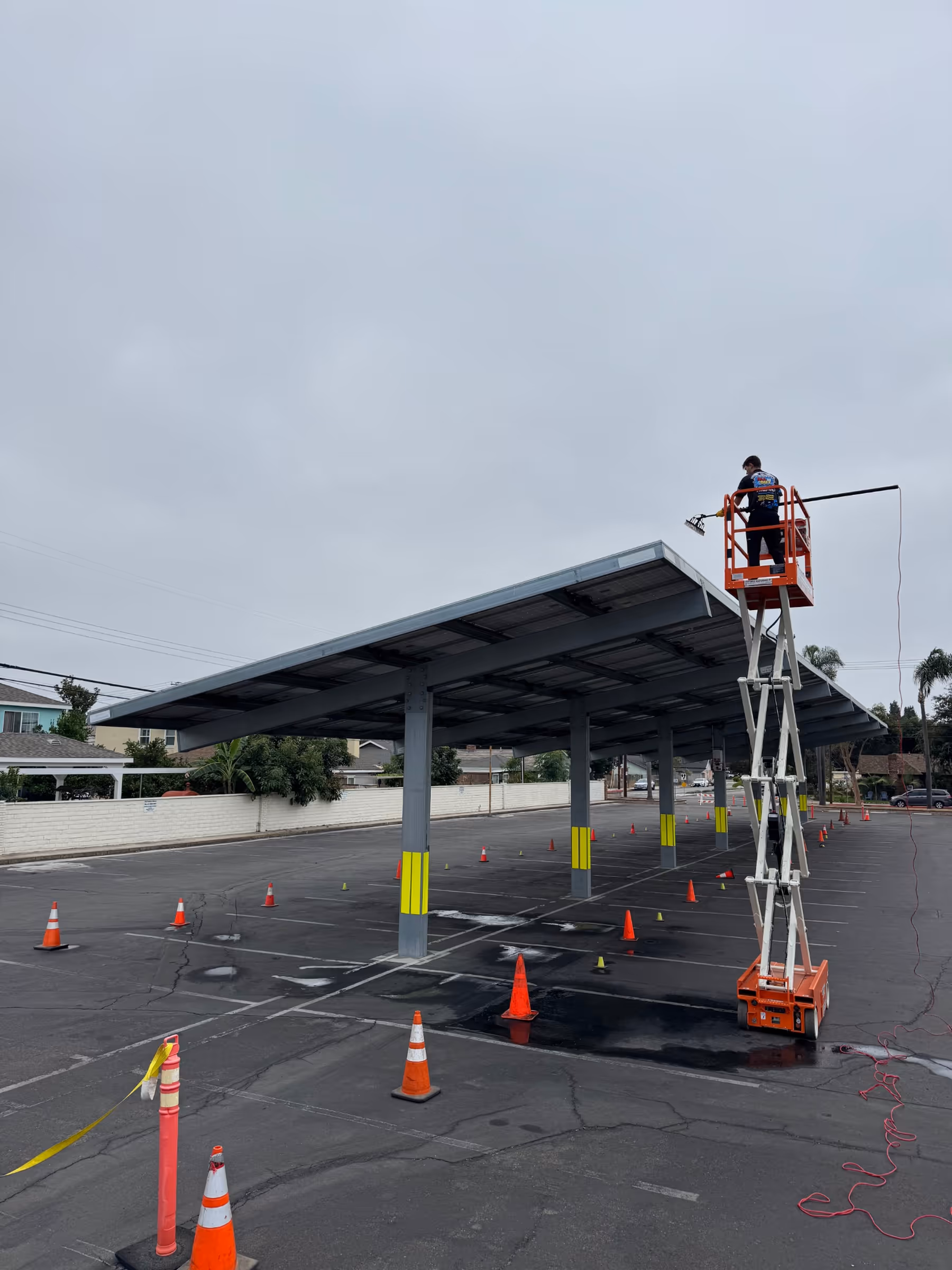 Worker using power washer to clean commercial solar canopy structure in parking lot.