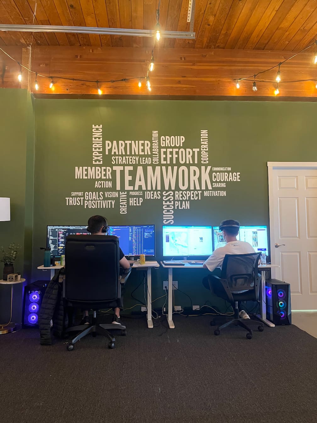 Two team members working at computer desks beneath a wall decorated with teamwork and collaboration words, highlighting a positive and supportive office environment.