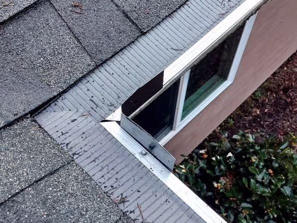 A close-up view of a house's roof corner with mesh gutter guards installed. The roof shingles appear clean and well-maintained, and the mesh gutter guards are securely fitted, preventing leaves and debris from entering the gutter system. The surrounding area includes a window and a landscaped garden.