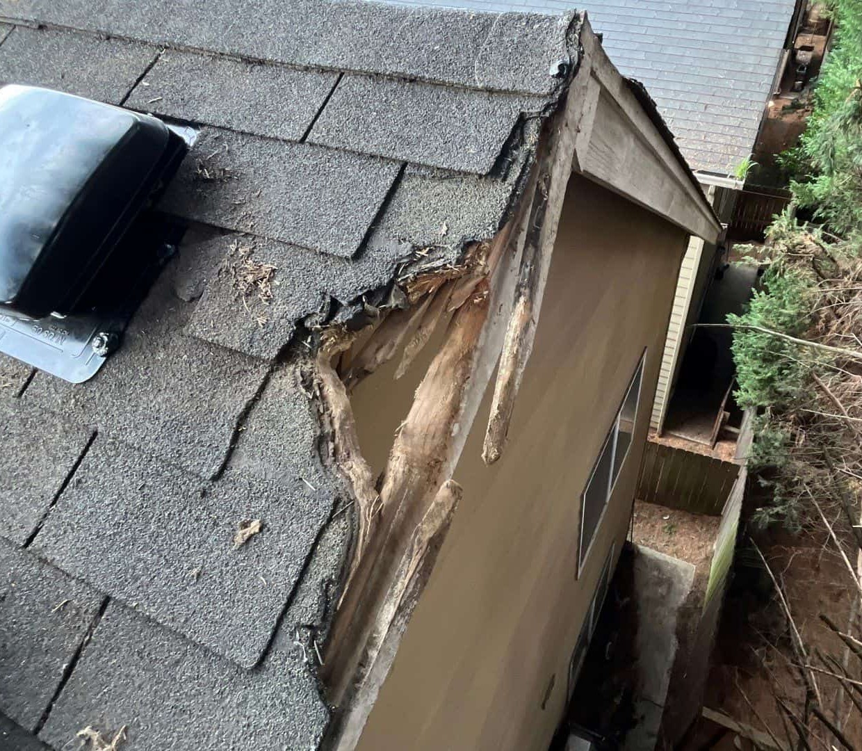 A close-up view of a roof corner severely damaged by a fallen tree branch. The shingles and wood structure are torn apart, exposing the underlying layers of the roof and fascia. Tree trunks and branches are visible beside the house, showing how the impact caused a large section of the roof to splinter and cave in.