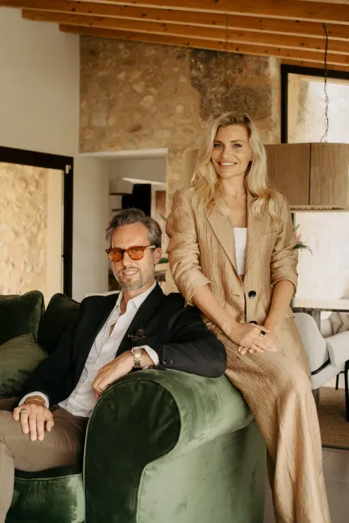 Stella and Elliot, the founders of By Costastay Property Management, pose in a stylish, modern interior. Stella stands beside the sofa, smiling and dressed in a light-colored suit, while Elliot sits on a plush green sofa, wearing a black blazer and sunglasses. The background features warm, earthy tones with wooden and stone elements, adding to the sophisticated and welcoming atmosphere.
