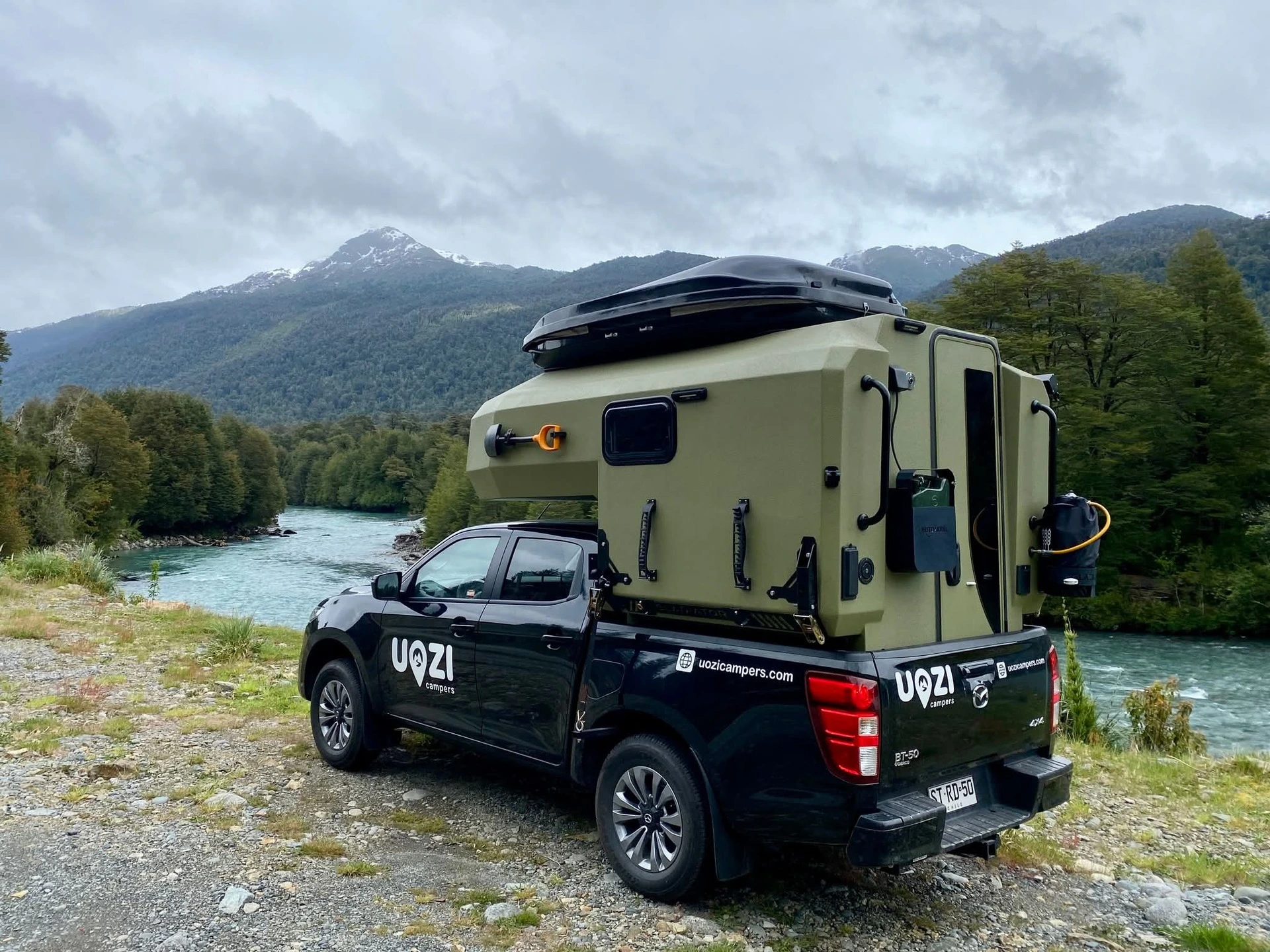 Green Uozi camper parked by a river between forest and mountains