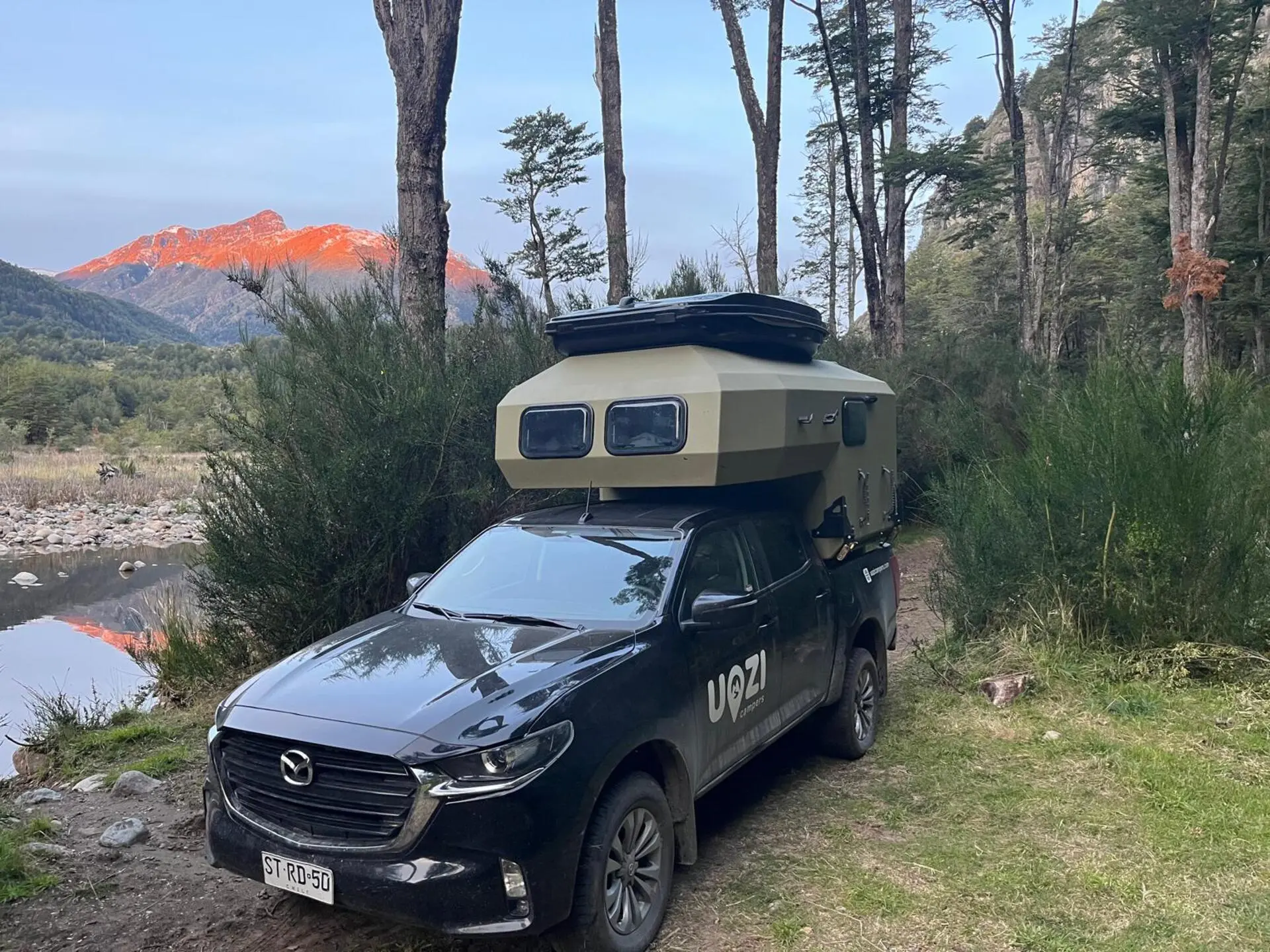 Uozi camper among trees with sunlit mountains at sunrise