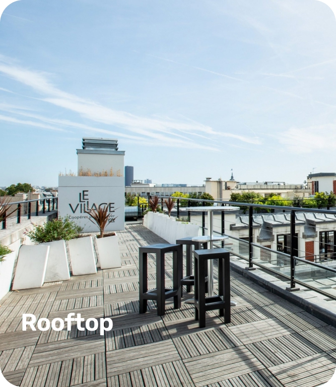 Modern rooftop terrace with wooden decking, black stools around a small table, potted plants, and a sign reading 'Le Village'.